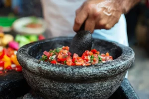 What is the point of a molcajete? 2 A hand using a tejolote to grind vegetables in a molcajete, surrounded by colorful ingredients.
