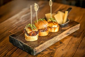 A wedge of smoked gouda with brisket sliders in the background, served on a wooden board.